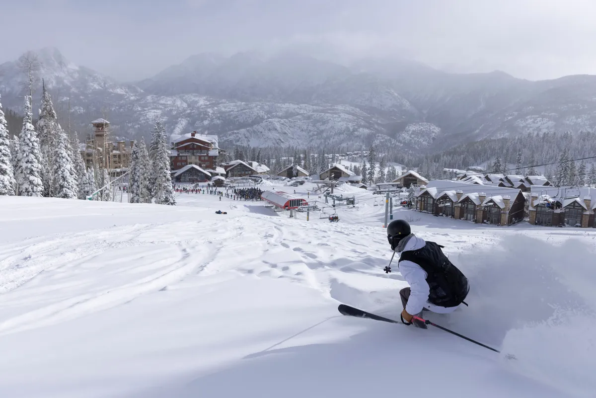 A dynamic exterior shot showing a skier in mid-action on a snow-covered slope, with Purgatory Resort buildings and snowy trees in the background, all framed by majestic mountains.