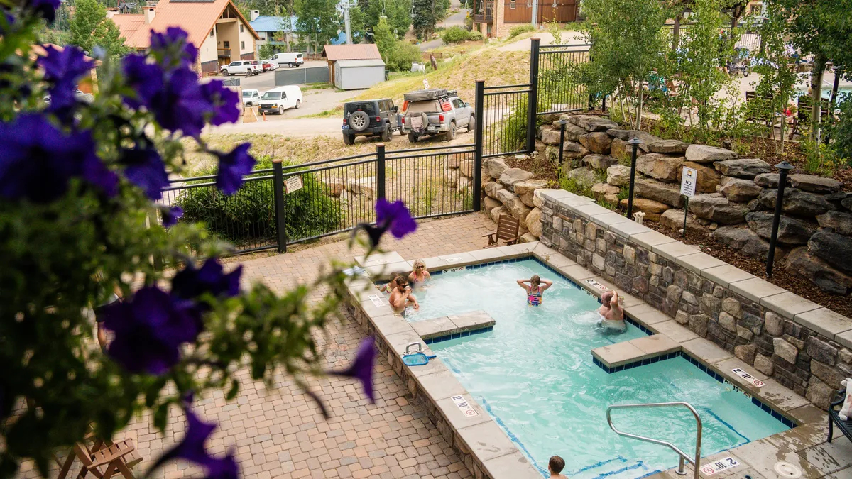 This image shows a sparkling outdoor swimming pool surrounded by lush greenery and colorful flowering plants. Several people are enjoying the water and relaxing on the pool deck, capturing the essence of a lively, resort-style amenity.