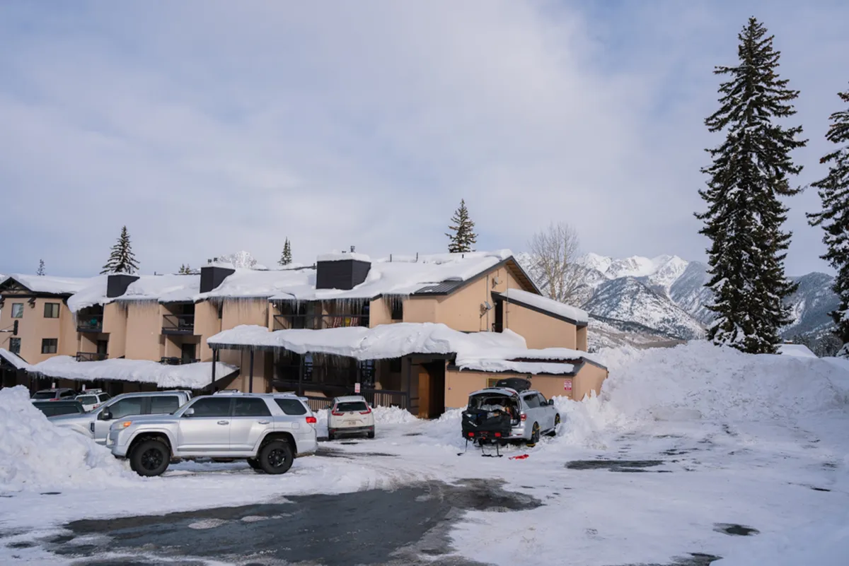 A sweeping exterior view featuring snow-covered roofs nestled at the base of the San Juan Mountains, with peaks rising in the background and hints of winter activity such as parked vehicles and subtle ski tracks. The tranquil winter landscape evokes a serene and inviting atmosphere, ideal for winter recreation enthusiasts.