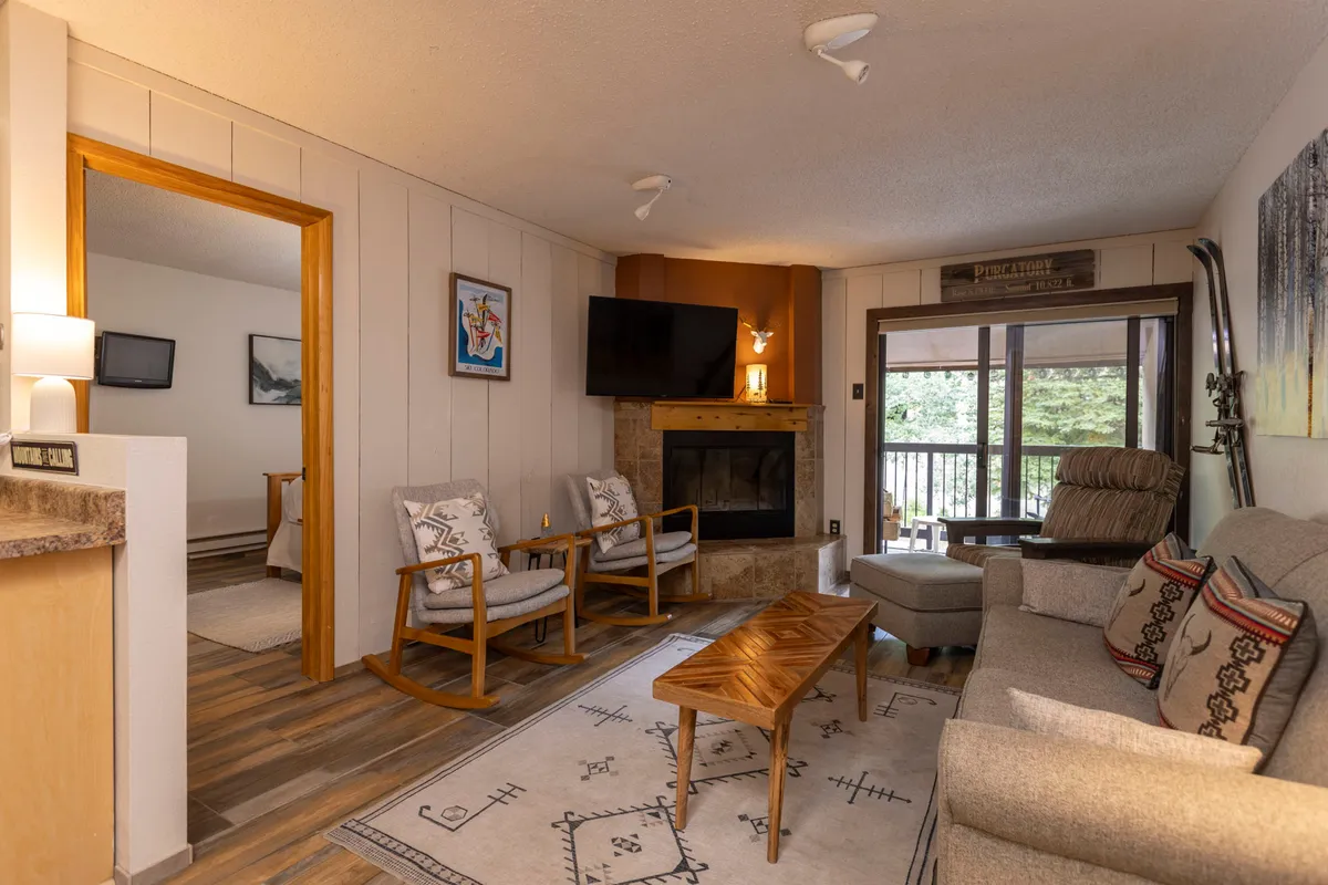 A cozy living room with a central fireplace, plush sleeper sofa, rocking chairs, and a coffee table. The room is bathed in warm light, encouraging guests to relax, socialize, or watch TV. Large windows are visible, letting in natural light and offering glimpses of the snowy alpine scenery outside.