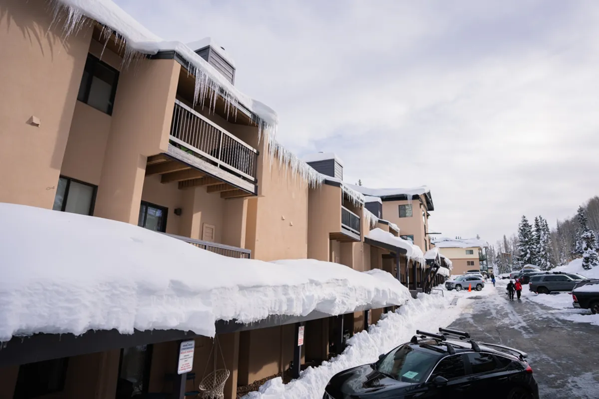 A charming view of Angel Haus's snow-covered roofs and balconies, with icicles hanging from the eaves and fresh snow blanketing the ground, capturing the inviting appeal of winter at the base of the mountain.