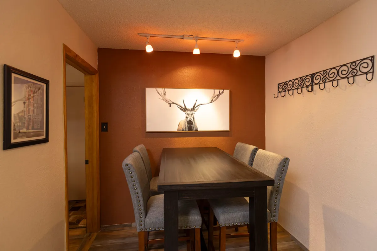 An inviting dining area featuring a large table, tasteful artwork, and warm decorative lighting. The table is set up for intimate meals, casual discussions, or games, and is positioned to benefit from the natural light streaming in.