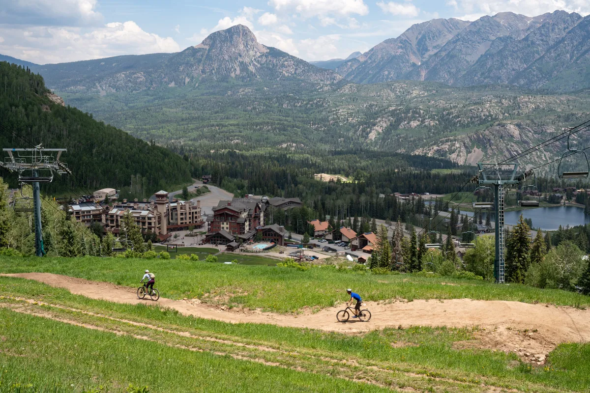 A wide view of the resort grounds featuring mountains in the background, cyclists enjoying the outdoors, and visible resort buildings and a chairlift. The scene conveys a sense of adventure and activity, with guests relaxing after exploring the mountains.
