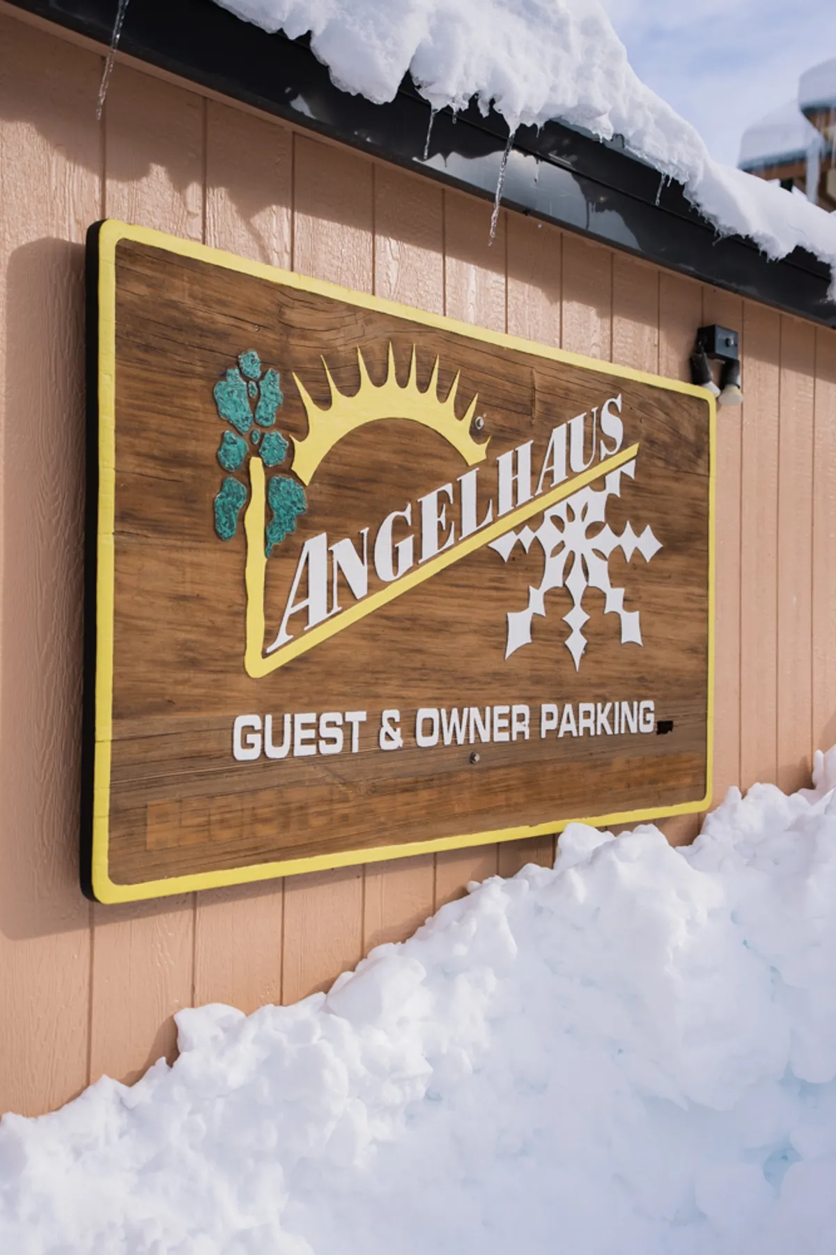 An inviting wooden sign stands amid a snow-covered ground, adorned with playful sun and snowflake graphics. The scene hints at easy access to both winter and summer activities, reinforcing the property's welcoming, year-round spirit.