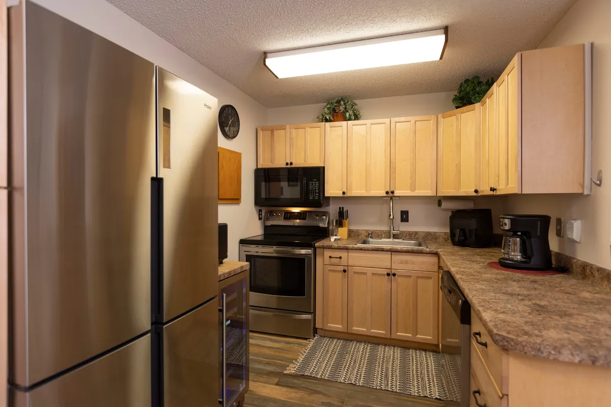 A modern kitchen scene with stainless steel refrigerator, ample wooden cabinetry, a well-lit countertop, and a sink with faucet. Soft lighting and thoughtful design create a cozy, welcoming atmosphere ideal for family gatherings and meal preparation.