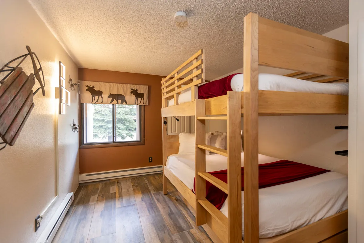 A family-friendly bedroom showcasing a unique queen-over-queen bunk bed, with playful curtains featuring a moose design on the window. The wooden flooring and relaxed decor invite families or groups to unwind and create memories, while the setup easily accommodates both kids and adults.