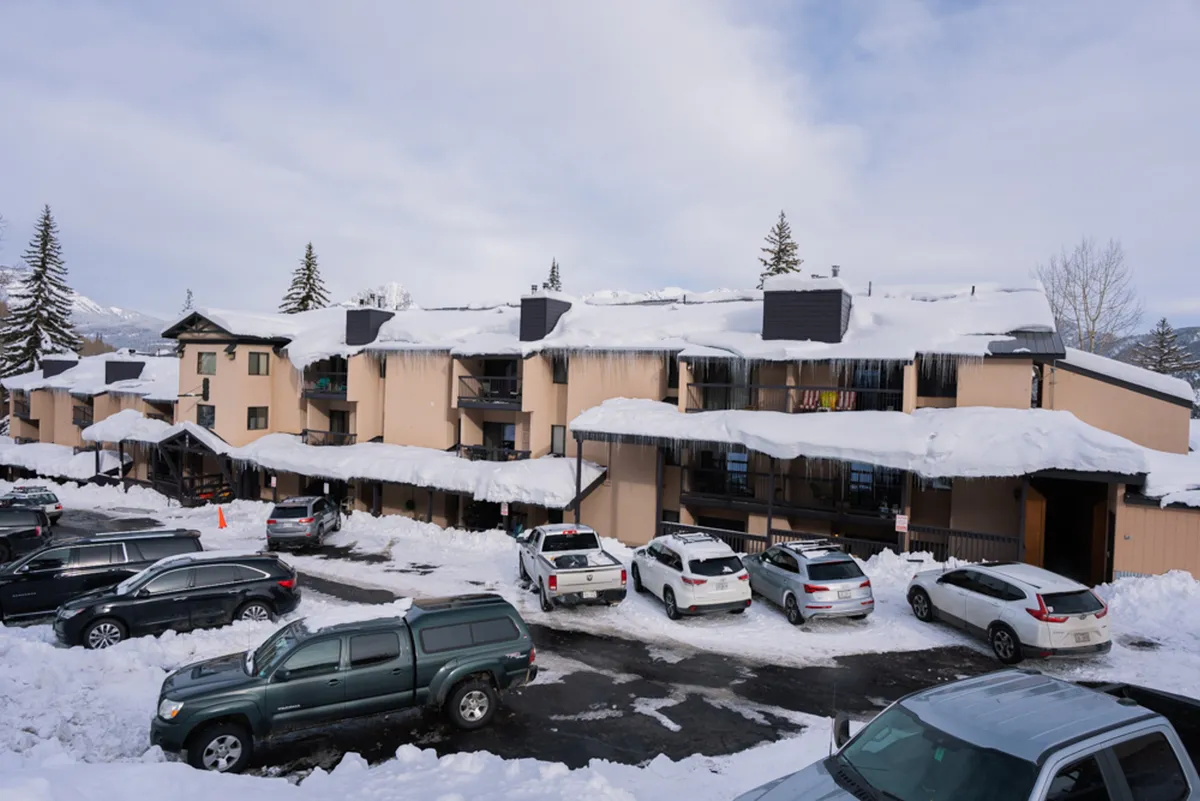 This exterior shot displays the snow-covered roofs of Angel Haus and nearby parked vehicles, framed by tall trees in the background. The scene invites thoughts of winter activities and the welcoming ambiance of the resort after a day on the slopes.